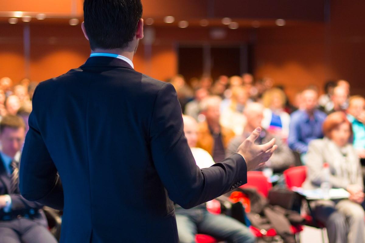A male figure with his back to us speaking to a crowded room